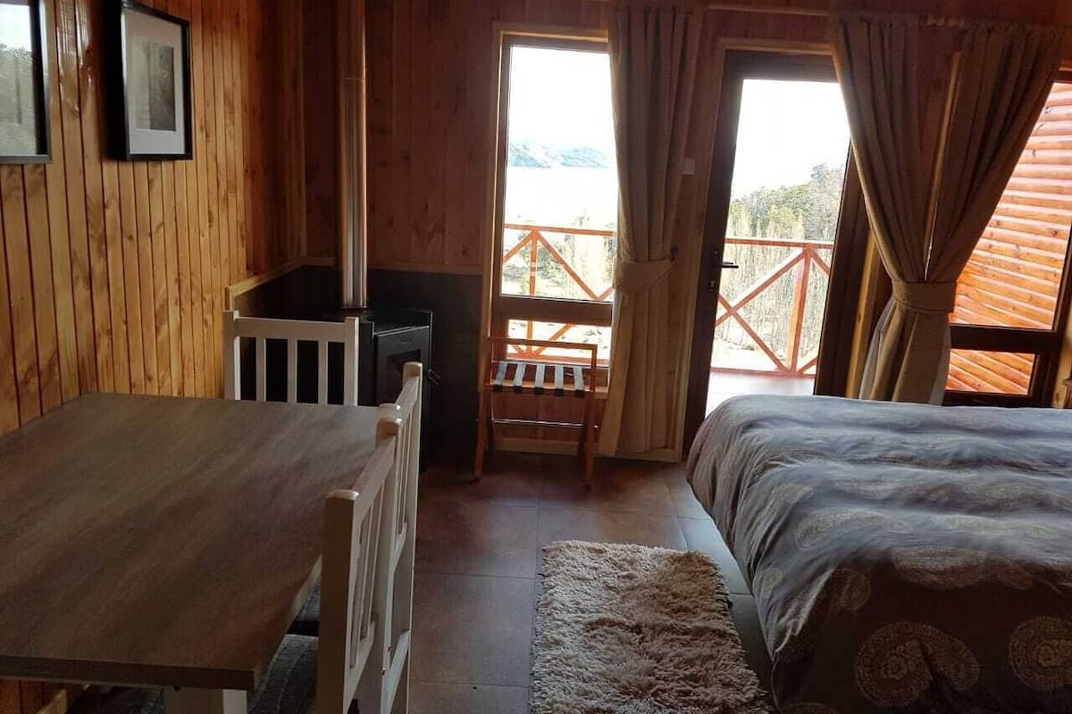 A wooden cabin room with a bed, a table, and a chair. There is a window with curtains and a view of a balcony.