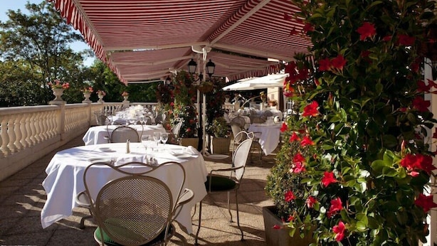 Un espace de restauration en plein air, avec des tables et des chaises placées sous des auvents à rayures, entouré de verdure et de plantes en pot.