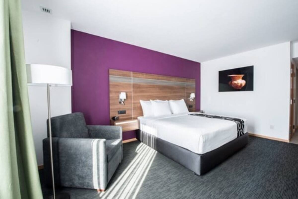 A hotel room with a bed, a chair, a nightstand, a lamp, and a wall-mounted picture.