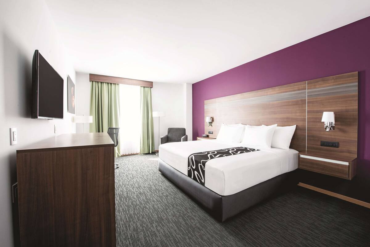 A modern hotel room with a large bed, a desk, a flat-screen TV, and a purple accent wall.