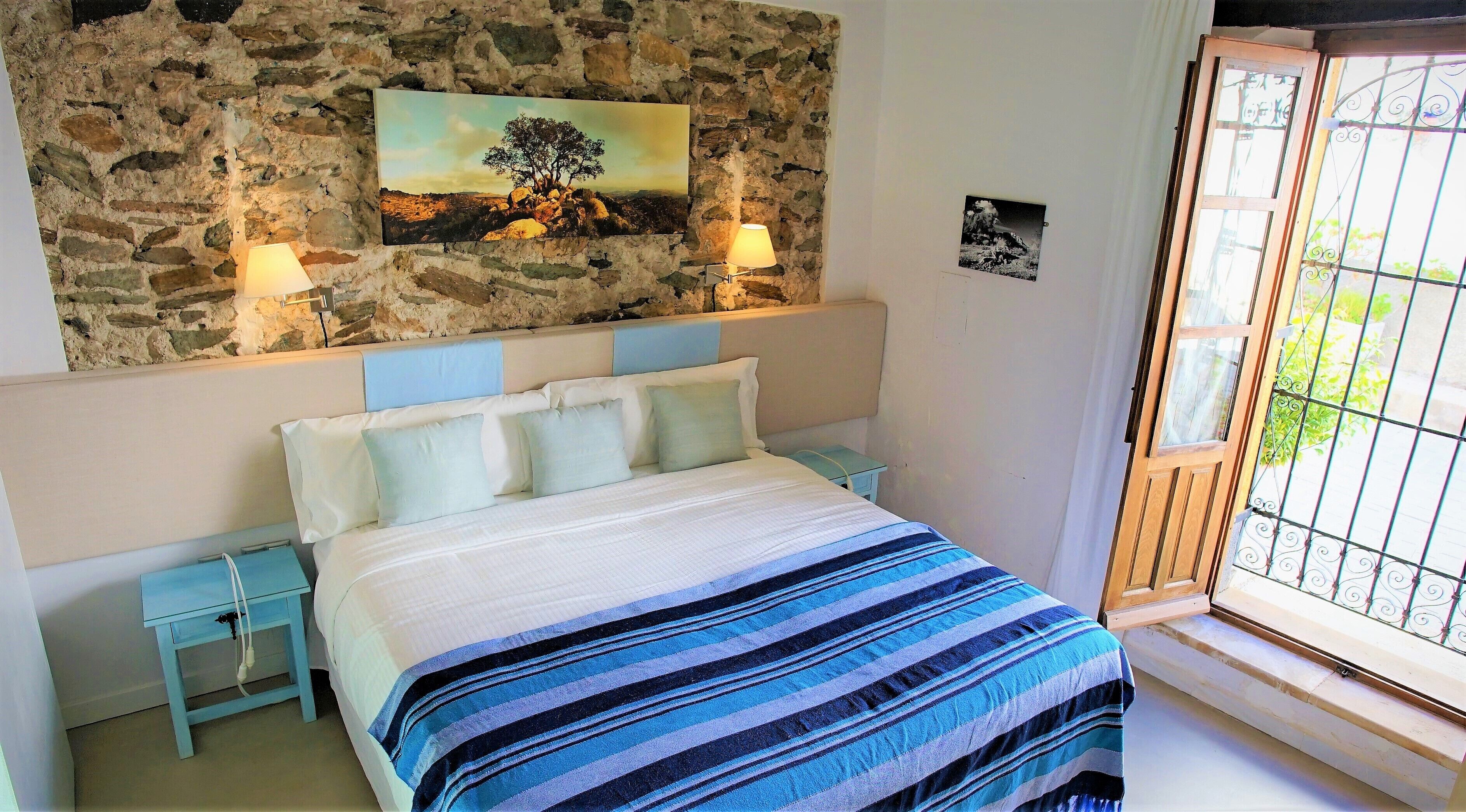 Un dormitorio con pared de piedra, una cama con manta a rayas, dos mesitas de noche, una lámpara y una ventana con vistas.