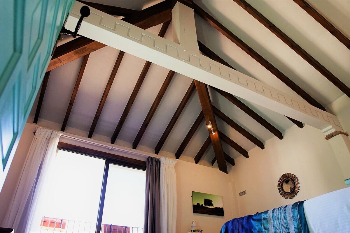 Una habitación con vigas de madera a la vista, una ventana con cortinas, un cuadro colgado en la pared y una cama con una manta azul.