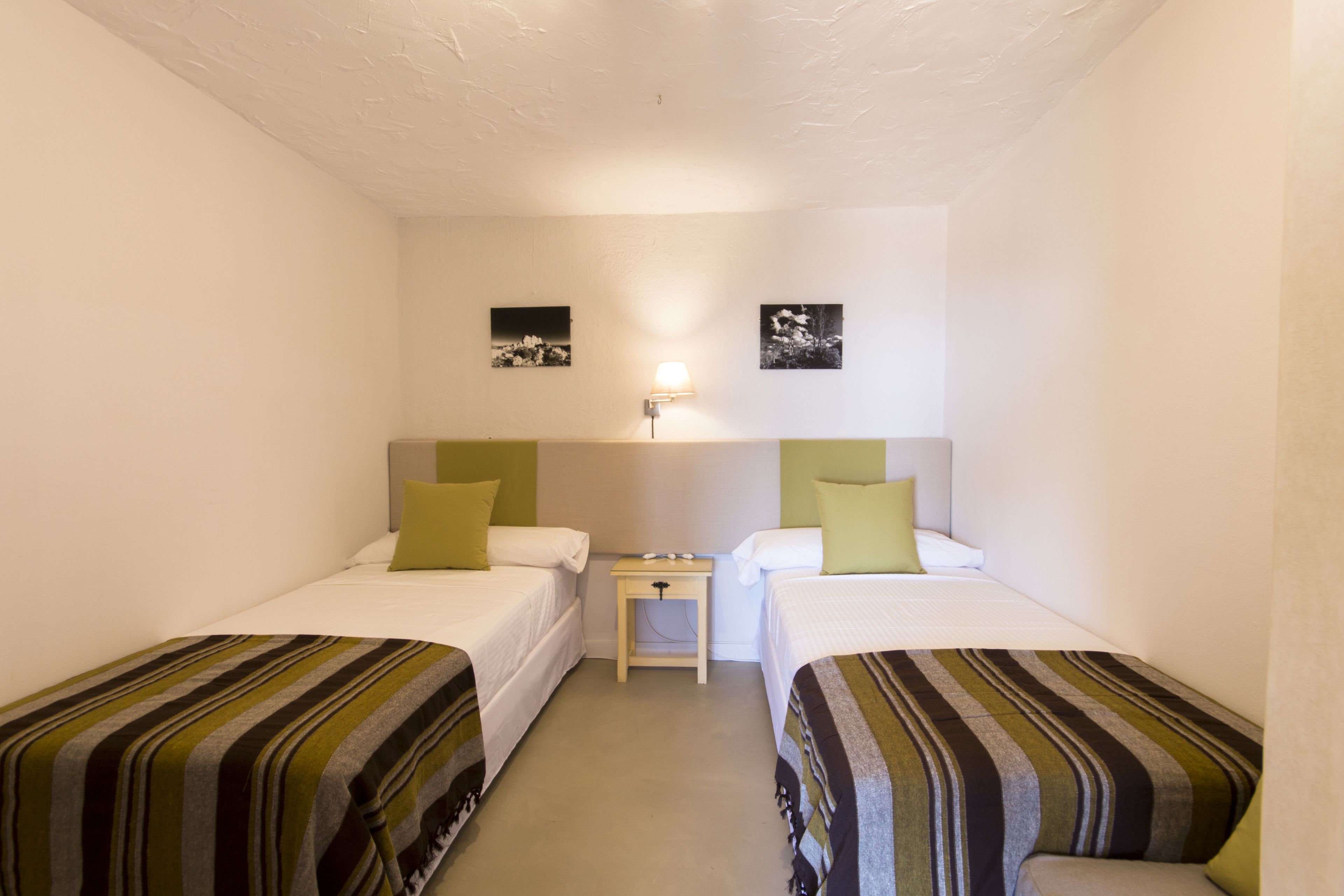 Une petite chambre propre, avec deux lits simples, une table de chevet et une lampe fixée au mur.