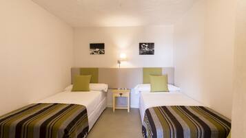 Une petite chambre propre, avec deux lits simples, une table de chevet et une lampe fixée au mur.