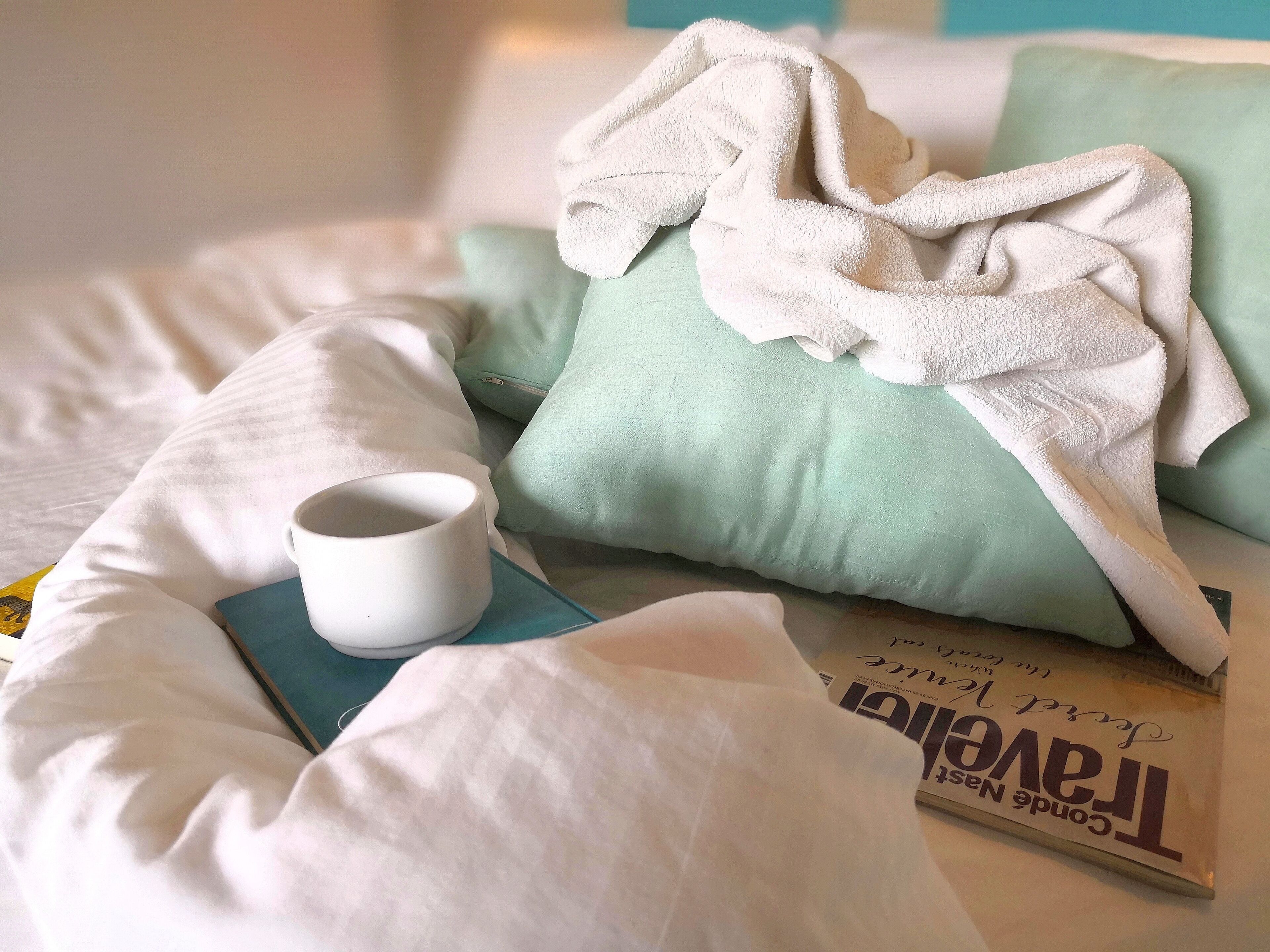 A neatly made bed with a white pillow, a teal pillow, and a white towel. There is a white mug on the bed and a book titled "The Great Gatsby" beside it.