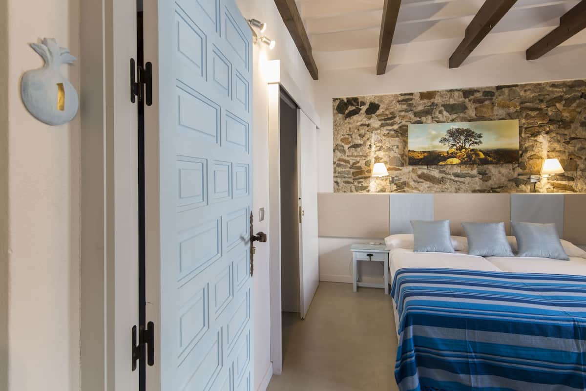 Un dormitorio con cama, mesitas de noche, una pared de piedra con un cuadro y una puerta blanca.