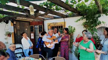 Un grupo de personas reunidas alrededor de una mesa con un guitarrista, en un espacio al aire libre techado con vigas de madera y vegetación.