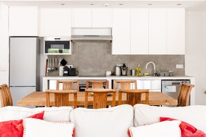 A modern kitchen with white cabinets, a built-in oven, and a dishwasher. There is a dining area with a wooden table and chairs. - Casa Das Cegonhas (Alcacer do Sal)