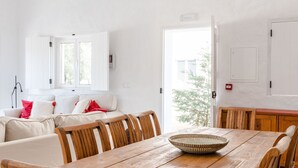 Ein Essbereich mit Holztisch und Stühlen, einem weißen Sofa mit roten Kissen sowie Blick ins Freie durch eine Tür.
