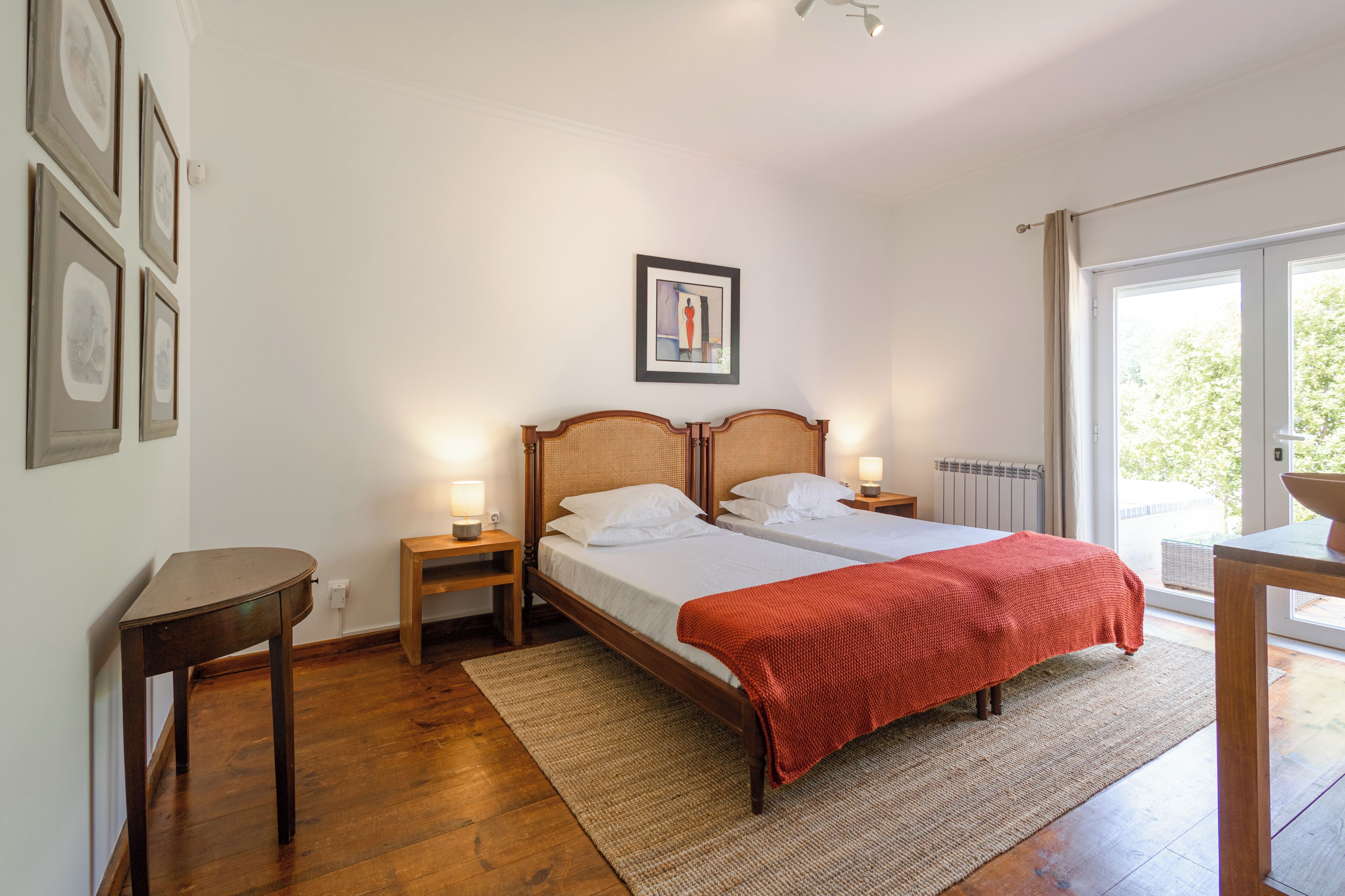 Un lit double avec une couverture rouge, deux tables de chevet avec des lampes, une petite table en bois, un tableau encadré au mur et, par la fenêtre, une vue sur la verdure.
