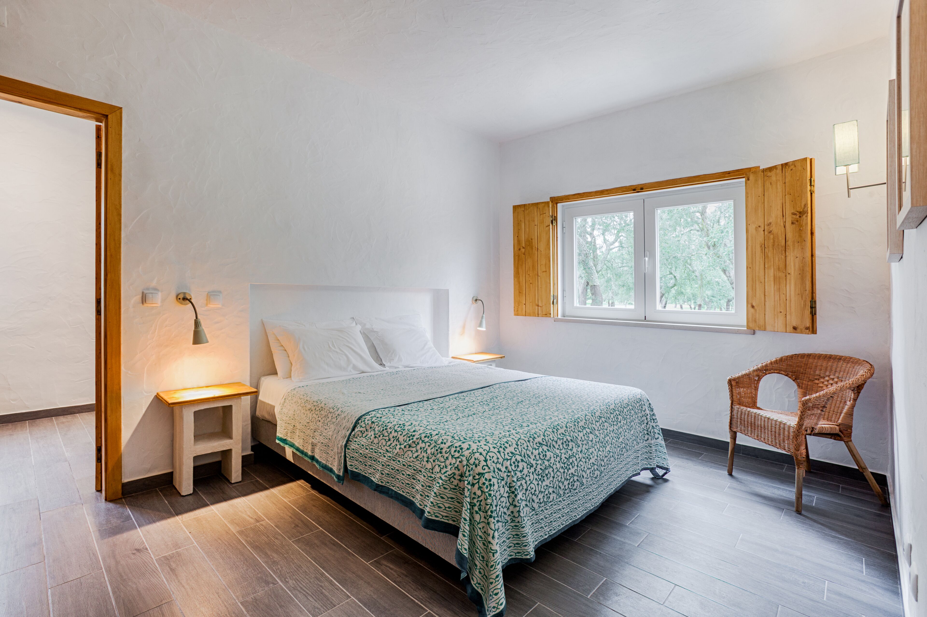 Ein Schlafzimmer mit Bett, Nachttisch, Lampe, Fenster und einem Korbstuhl.