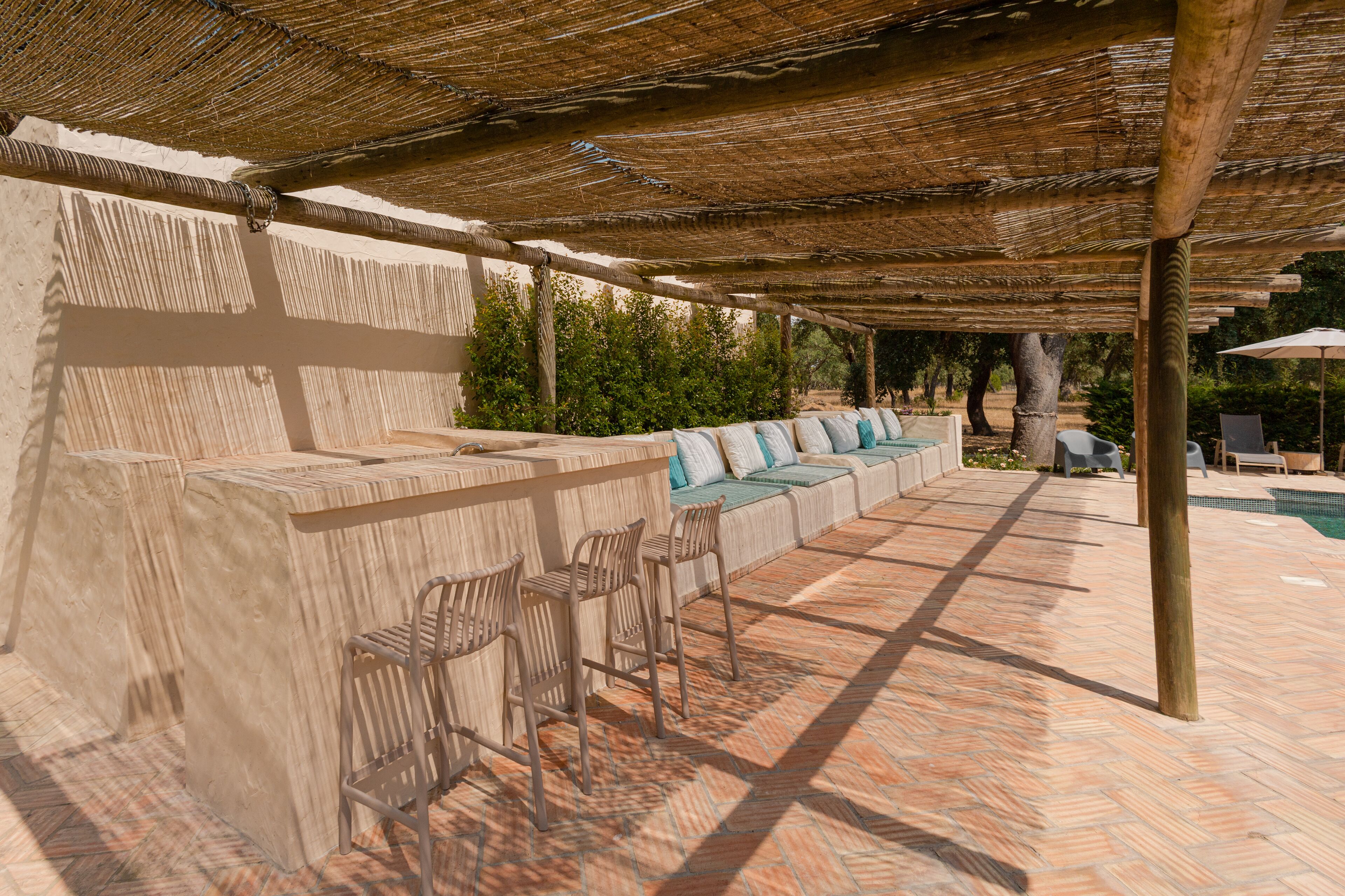 An outdoor seating area with a thatched roof, stone benches, and a pool in the background.