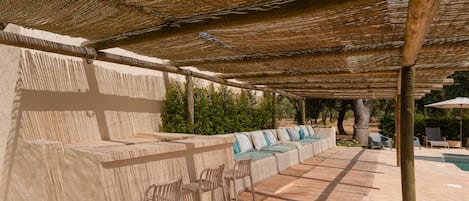 An outdoor seating area with a thatched roof, stone benches, and a pool in the background.