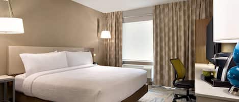 Habitación de hotel con una cama grande, un escritorio con lámpara, una silla y un ventanal con cortinas.