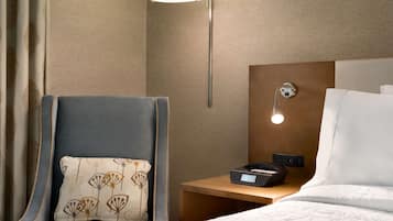 Habitación de hotel con cama, mesita de noche con lámpara, silla con cojín estampado y teléfono en la pared.