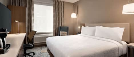 Quarto de hotel com uma cama grande, uma escrivaninha com cadeira, abajur e janela com cortinas.