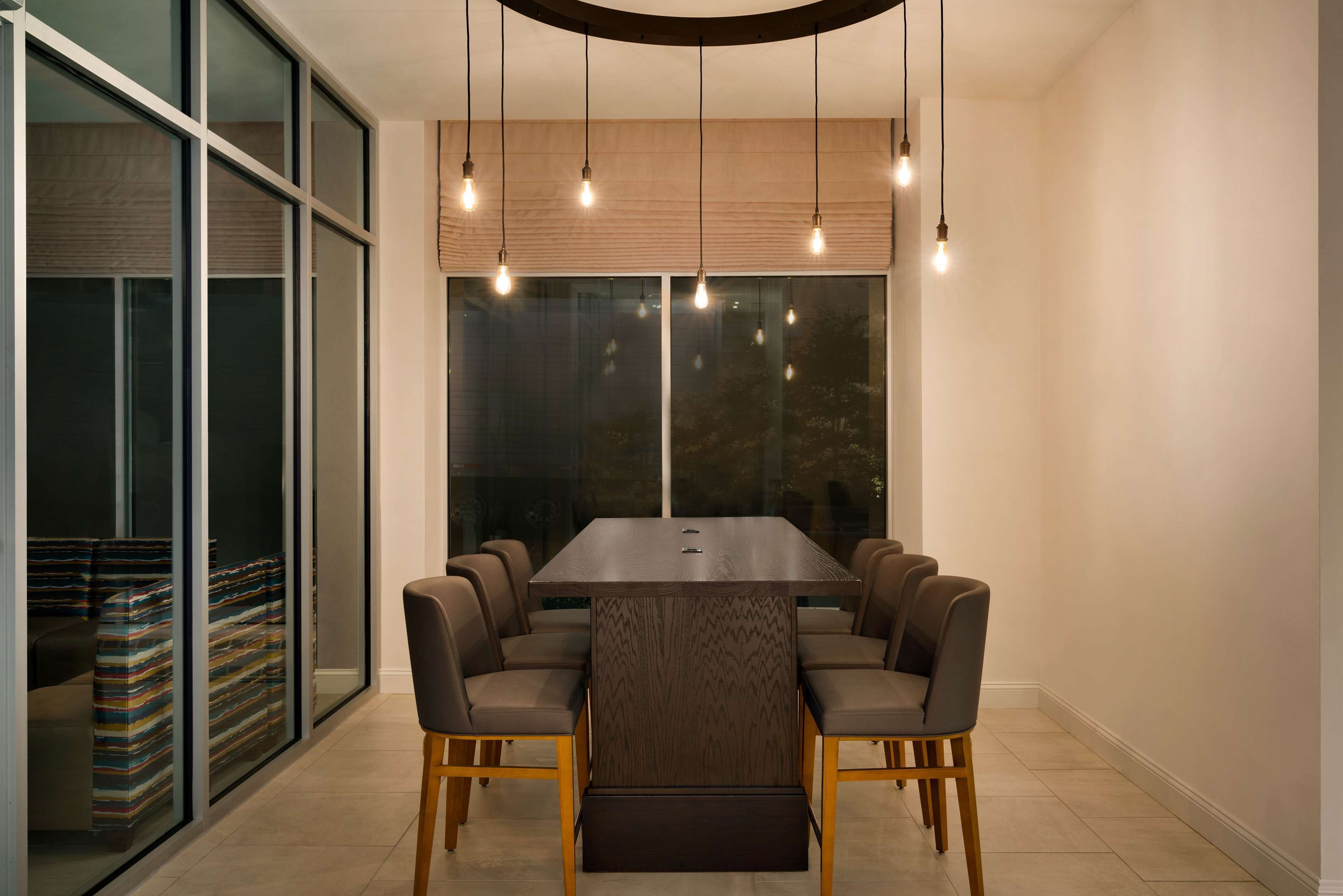 A modern dining area with a rectangular table surrounded by six chairs, large windows with blinds, and hanging pendant lights.
