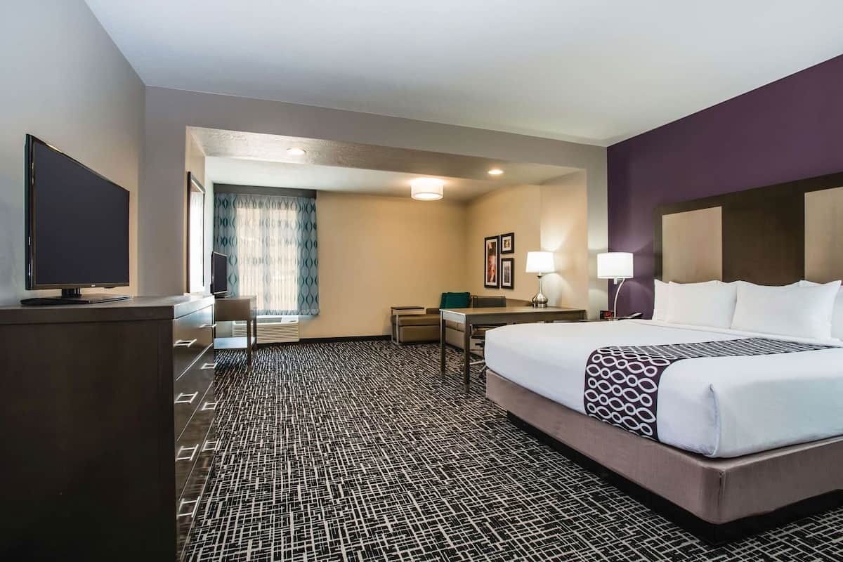 Ein Hotelzimmer mit einem großen Bett, einem Schreibtisch, einem Sessel, einem Fernseher und einem Fenster mit Vorhängen.