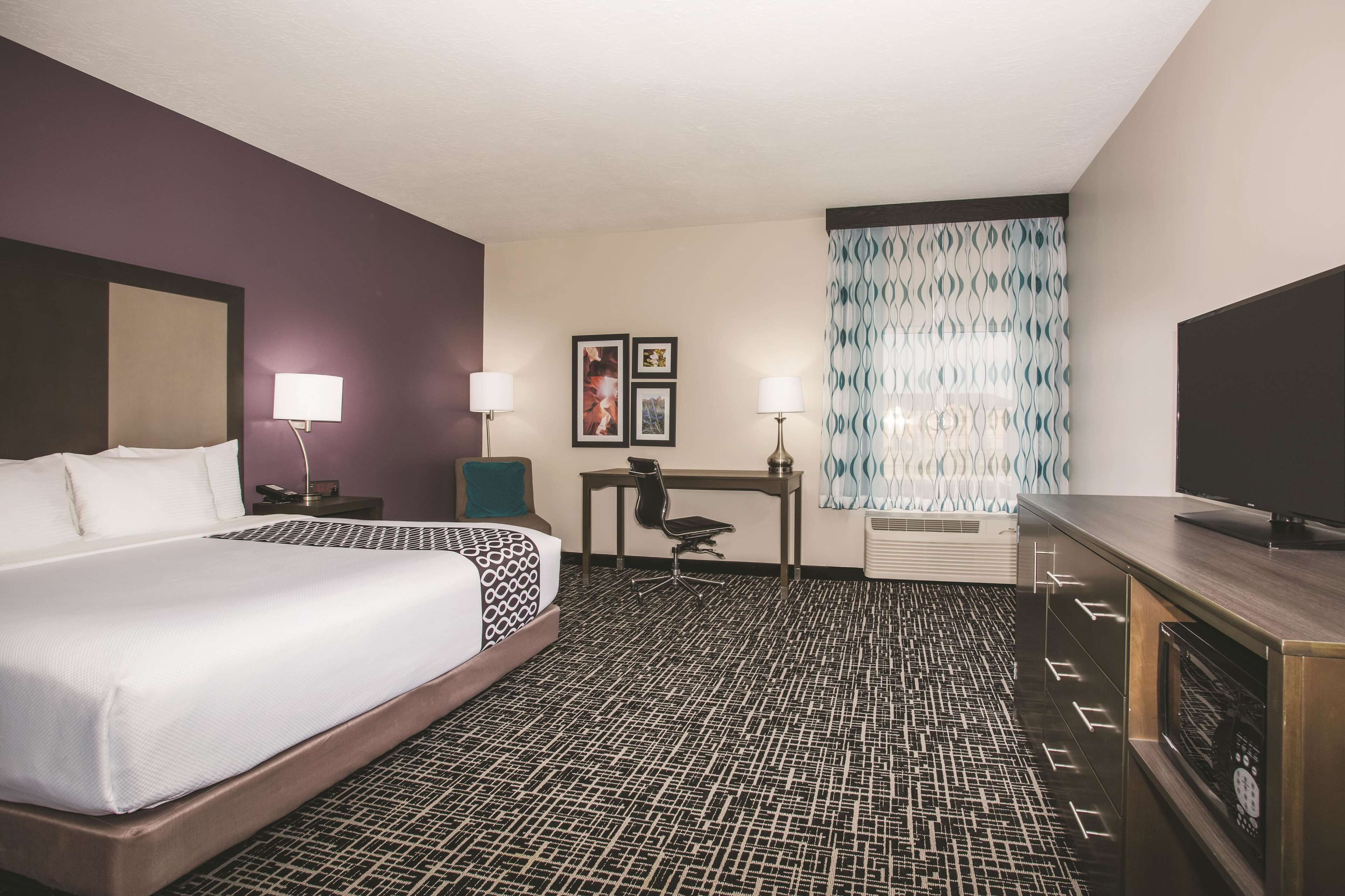 A hotel room with a large bed, a desk, two chairs, a television, and a patterned carpet.