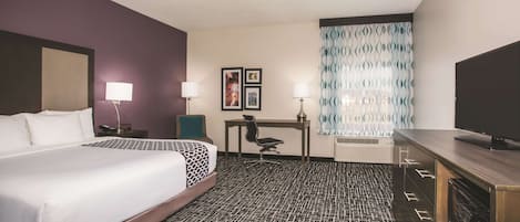 A hotel room with a large bed, a desk, two chairs, a television, and a patterned carpet.