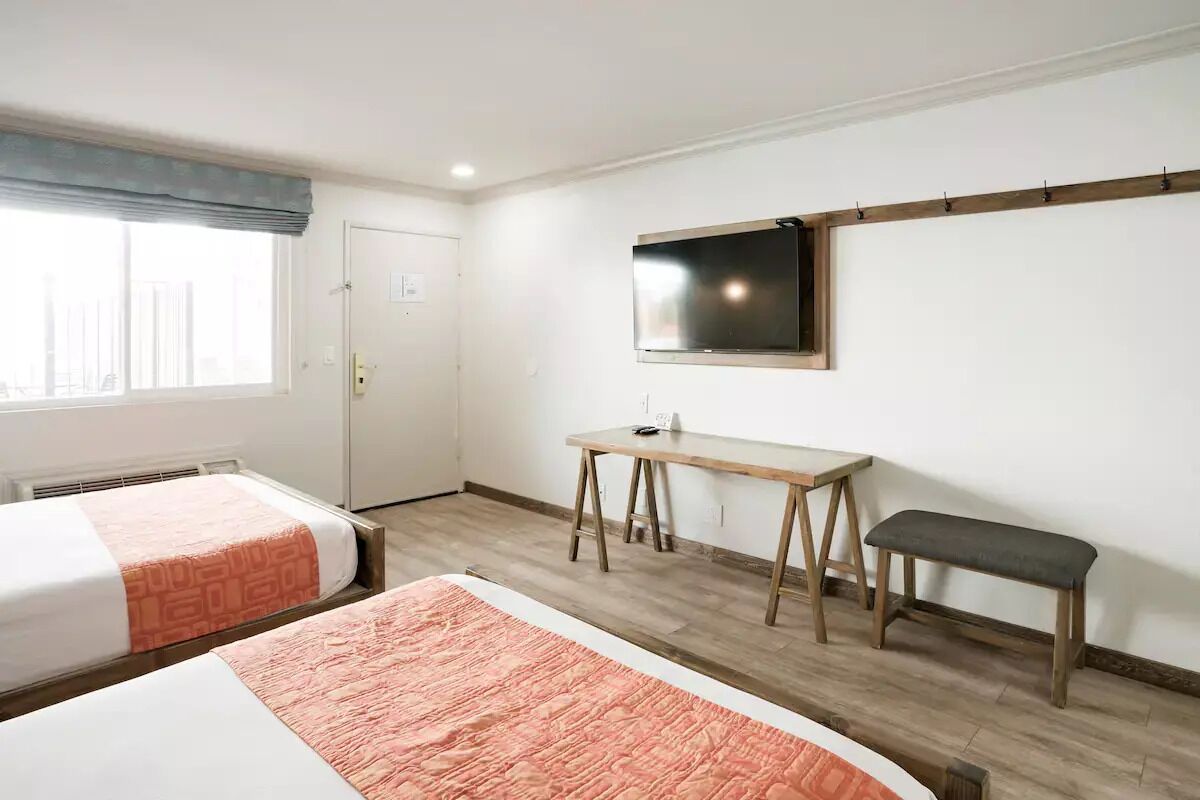 Ein Hotelzimmer mit zwei Betten, einem Schreibtisch, einer Bank, einem Fernseher und einem Fenster mit Vorhang.