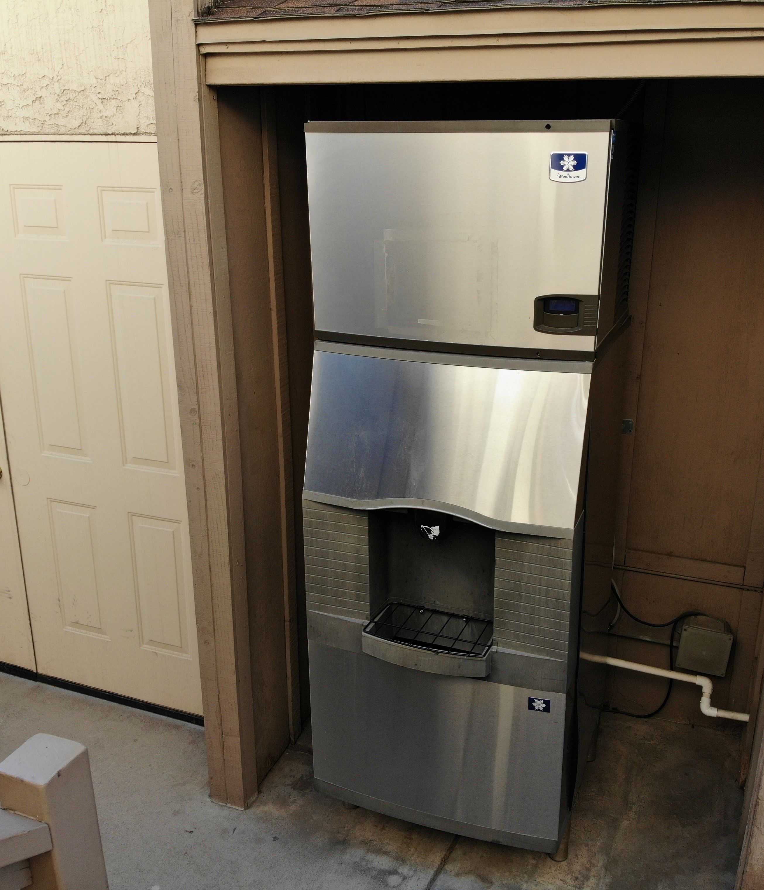 A stainless steel refrigerator with a water dispenser, located in a garage or utility area.