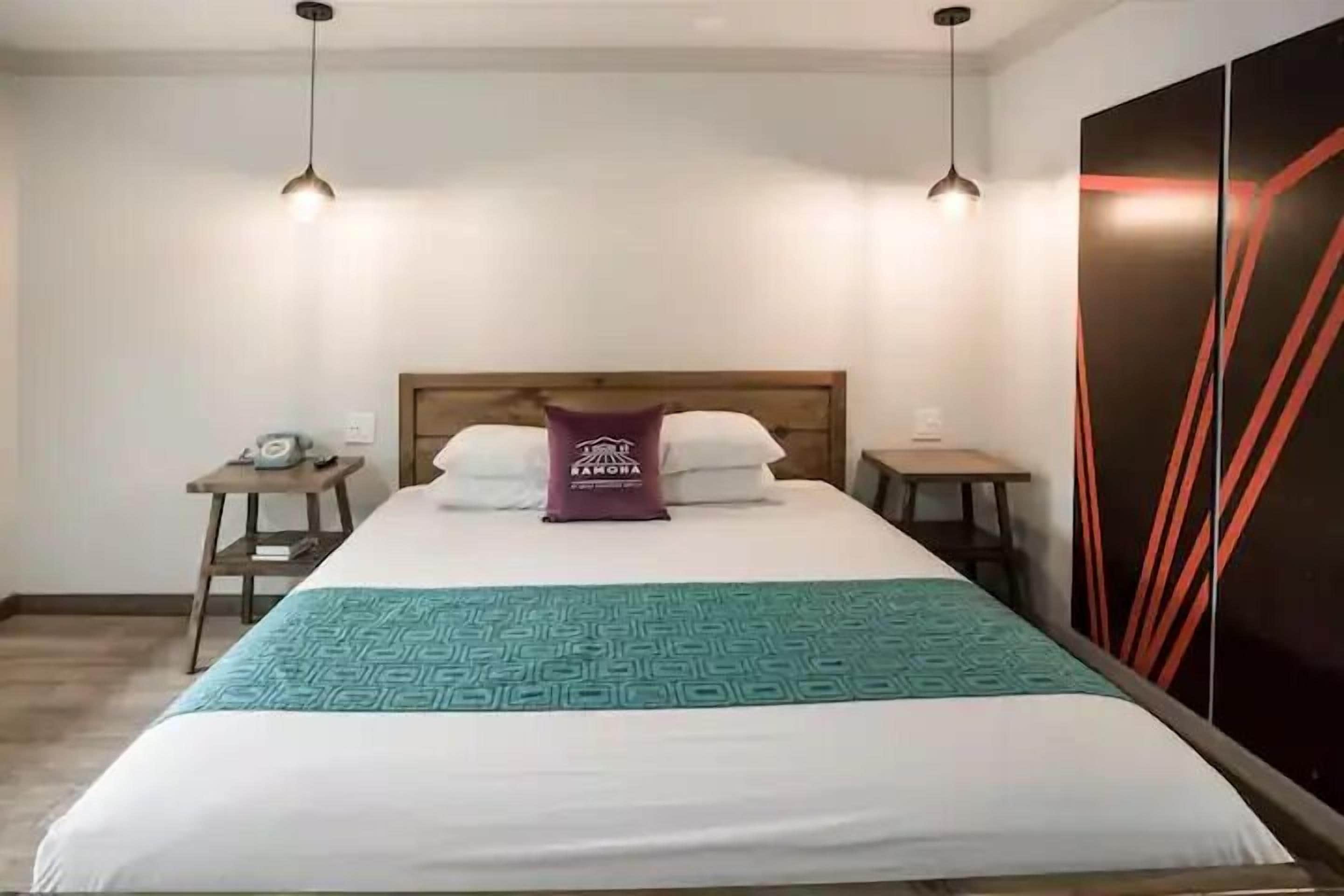 Una cama bien tendida con una colcha estampada, un almohadón con texto y dos mesitas de noche con lámparas.
