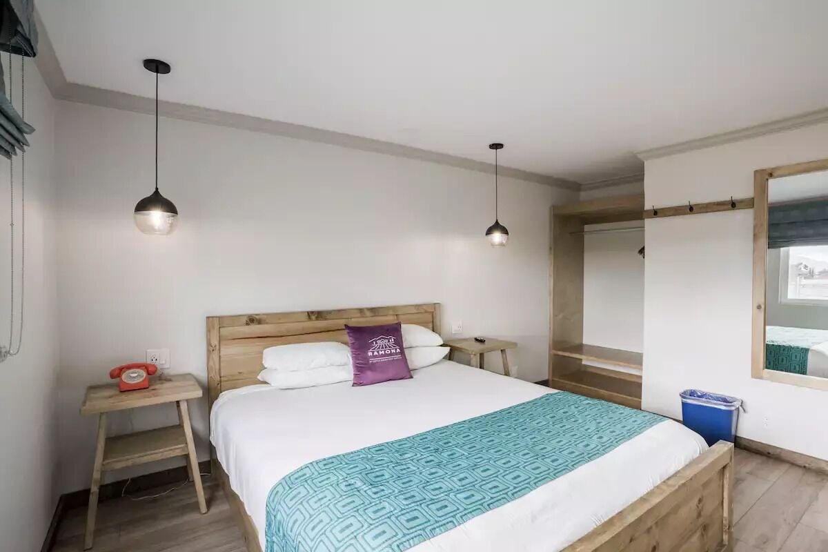 Een slaapkamer met een houten bed, twee nachtkastjes, een rode telefoon, een paars kussen, een blauwe prullenbak en een raam met uitzicht.