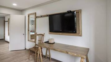 Una habitación con un escritorio de madera, una silla, un espejo grande y un televisor de pantalla plana montado en la pared.