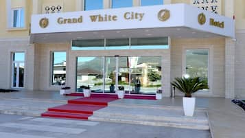 Eingang des Grand White City Hotels mit rotem Teppich, der zu den Türen führt, großen Fenstern und einem Blumentopf auf der rechten Seite.