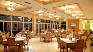 A dining area with multiple tables and chairs, a chandelier, and a television screen on the wall. - Grand White City (Velabisht)