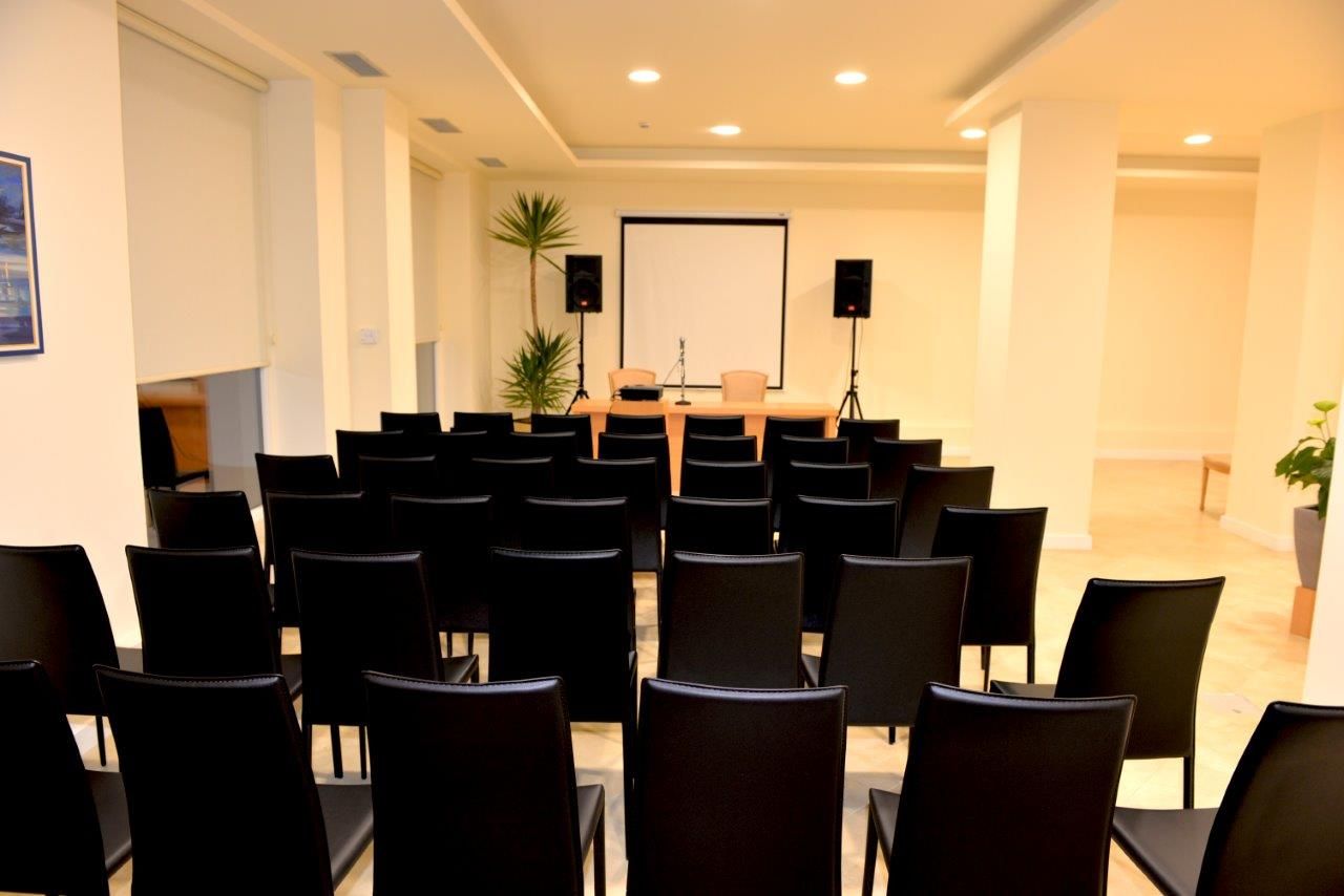 A conference room with rows of black chairs, a podium, a projector screen, and two speakers.