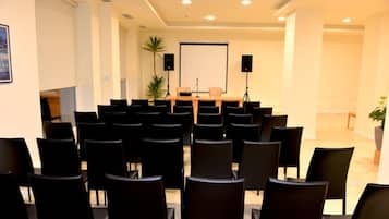 A conference room with rows of black chairs, a podium, a projector screen, and two speakers.