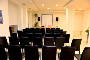 Een congreszaal met rijen zwarte stoelen, een spreekgestoelte, een projectiescherm en twee sprekers.