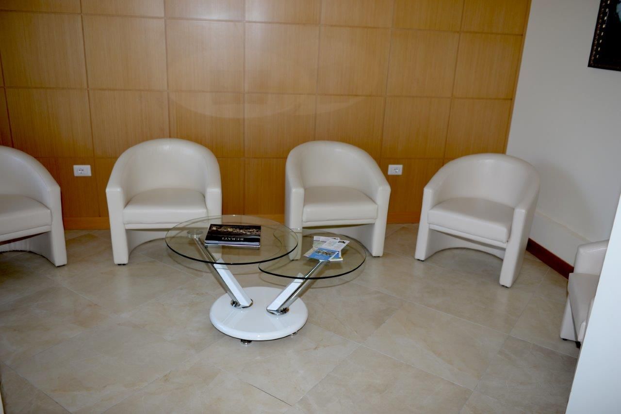 A waiting area with three white chairs, a glass-top table with a book and magazine, and a tiled floor.