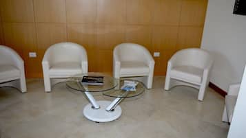 A waiting area with three white chairs, a glass-top table with a book and magazine, and a tiled floor.