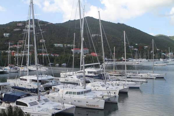Un puerto deportivo con varios veleros amarrados, rodeado de un paisaje montañoso y edificios al fondo.