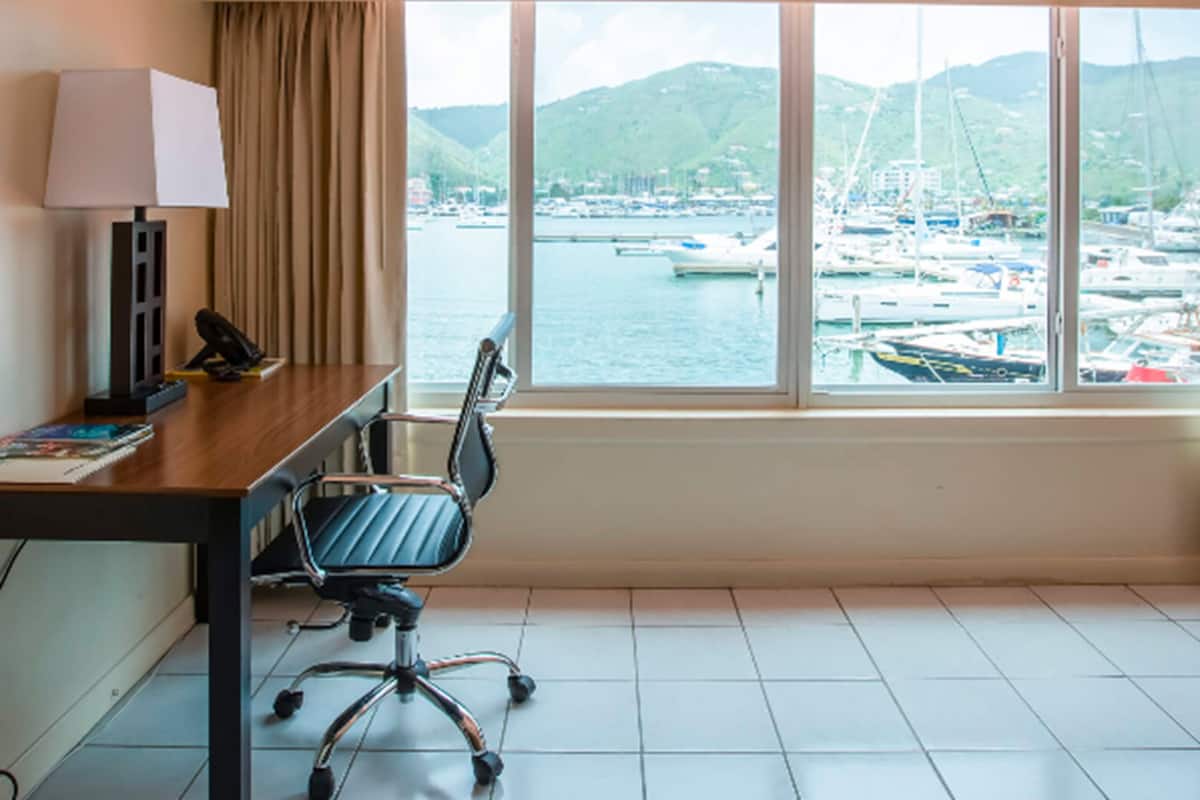 Een hotelkamer met een bureau, stoel, lamp en een groot raam met uitzicht op een jachthaven met boten en heuvels.