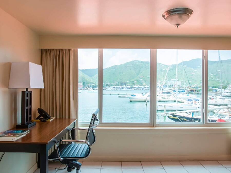 Quarto de hotel com escrivaninha, cadeira, luminária e uma ampla janela com vista para uma marina com barcos e colinas.