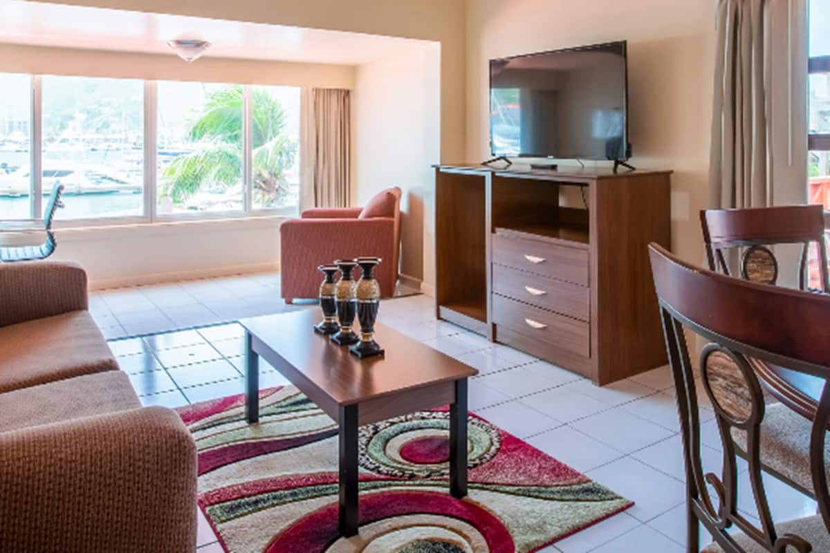 Een hotelkamer met een plafondventilator, een televisie op een houten standaard, een bank, een eettafel met stoelen en uitzicht op de jachthaven door het raam.