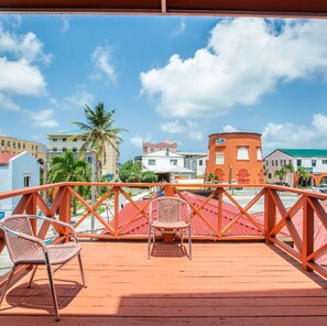 Una terraza de madera con dos sillas de mimbre, con vistas a un pueblo costero con edificios y palmeras.