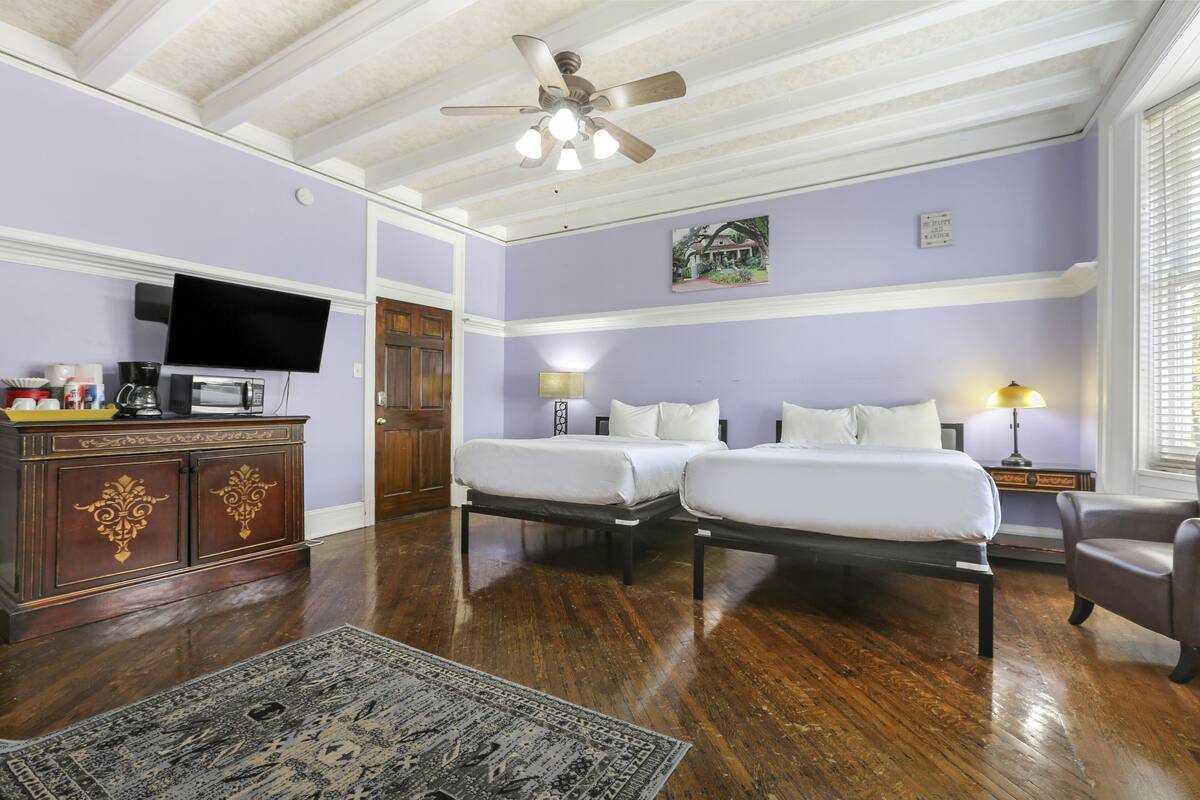 A hotel room with two beds, a television, a desk with a computer, and a ceiling fan.