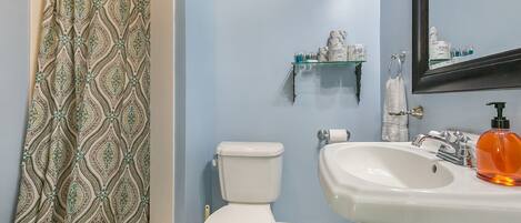 Une salle de bain avec un rideau de douche à motifs, des toilettes, un lavabo avec un distributeur de savon et une étagère contenant des articles de toilette.