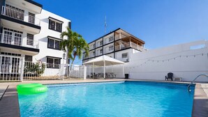 A swimming pool with a green inflatable ring, a covered seating area, and a white building with balconies.