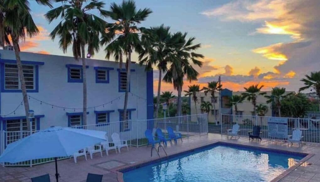 Una piscina con sillas de descanso y sombrillas, rodeada de palmeras y un edificio con detalles azules.