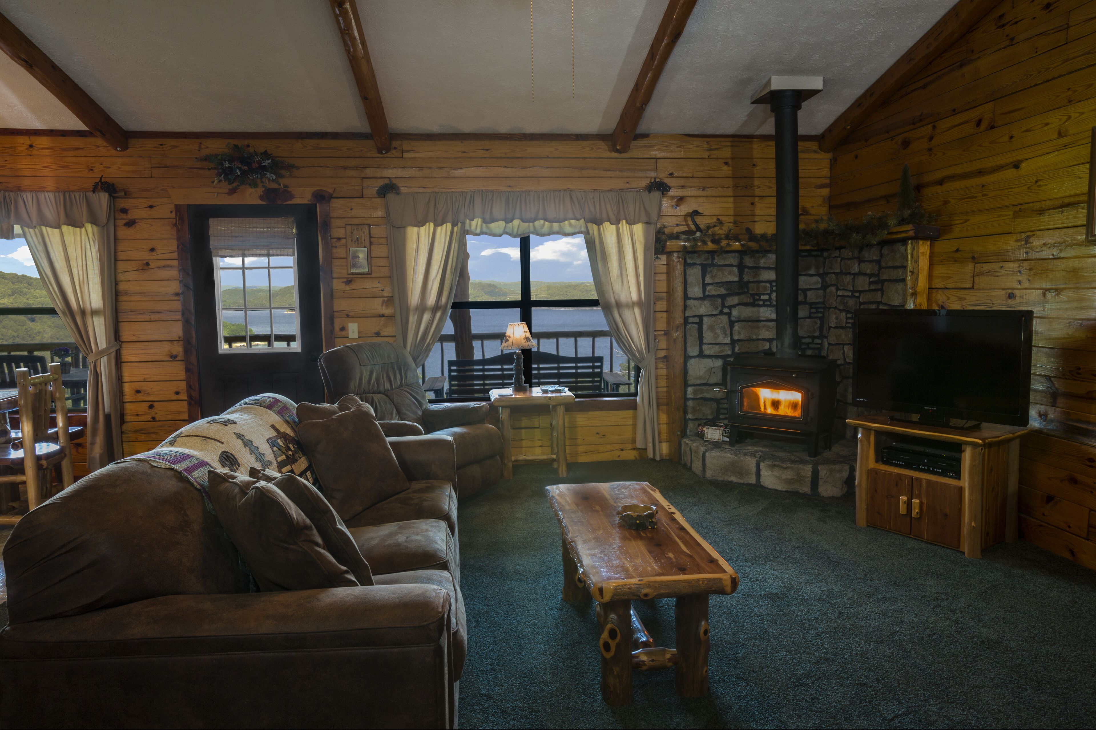 A cozy cabin interior with a stone fireplace, wooden furniture, and a large window overlooking a scenic landscape.