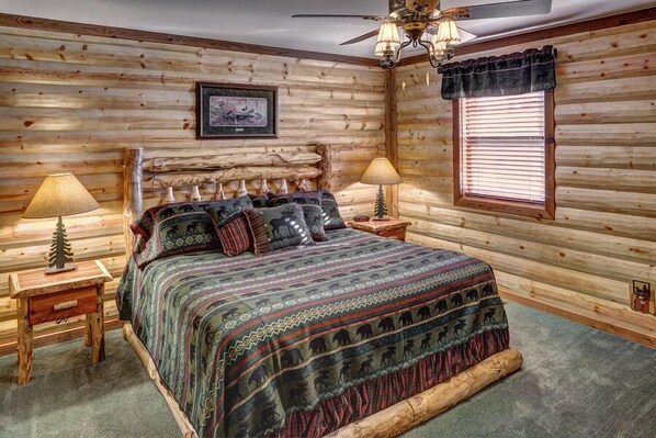 Um quarto no estilo de cabana de tronco, com cama, duas mesas de cabeceira, luminárias, um ventilador de teto e uma janela com persianas.