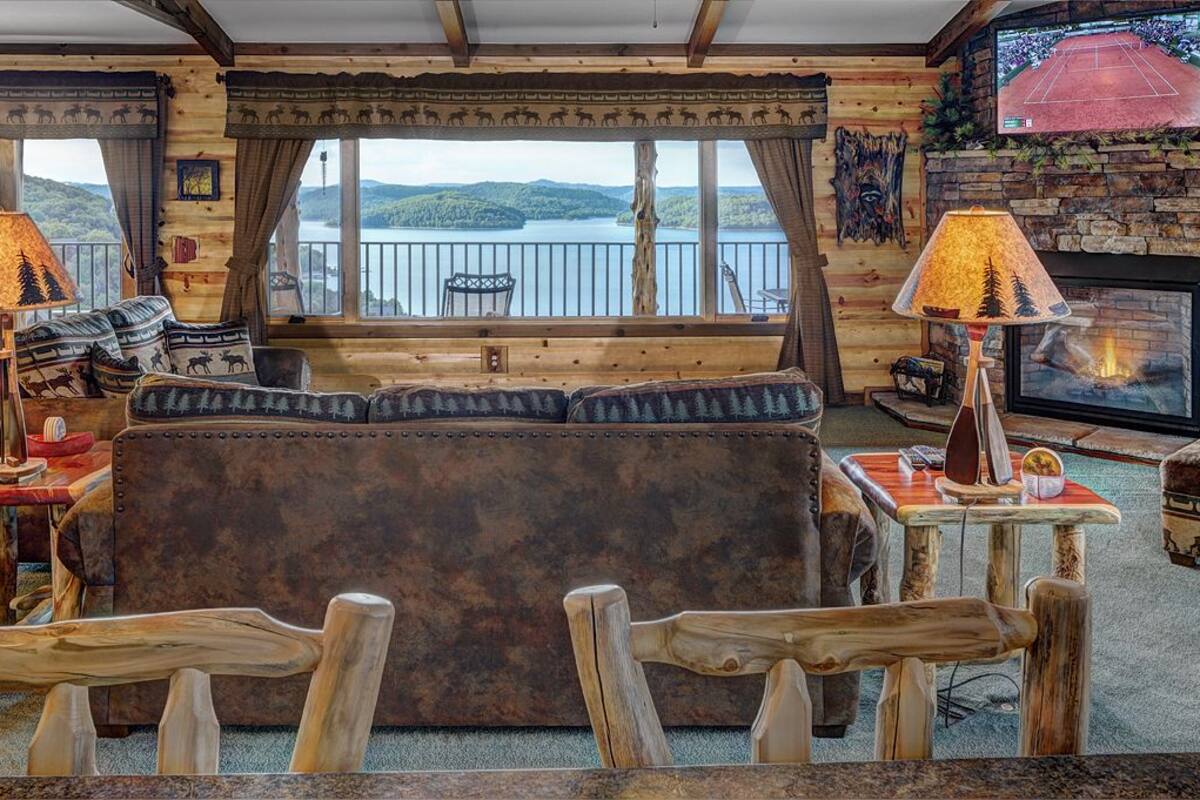 Un interior de cabaña acogedor con un gran ventanal con vistas a un lago, una chimenea y muebles de madera rústica.