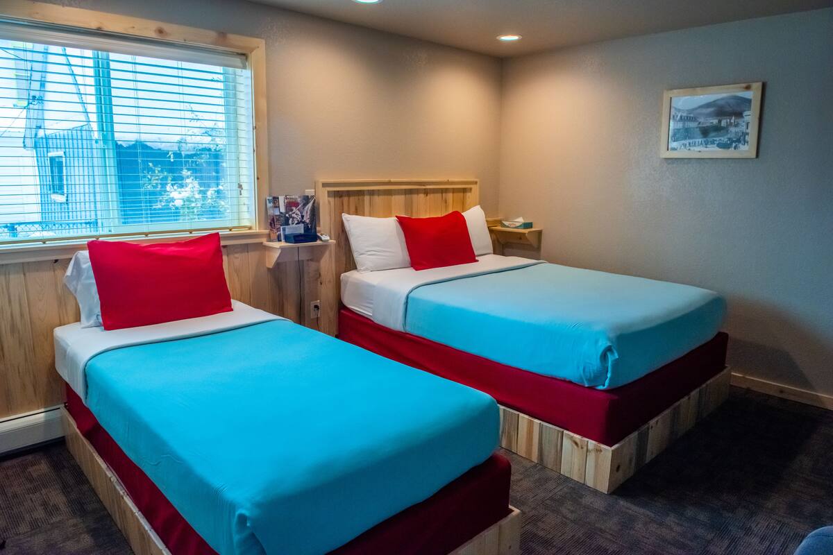 A room with two beds, each with a wooden headboard, white and red bedding, and a window with blinds.