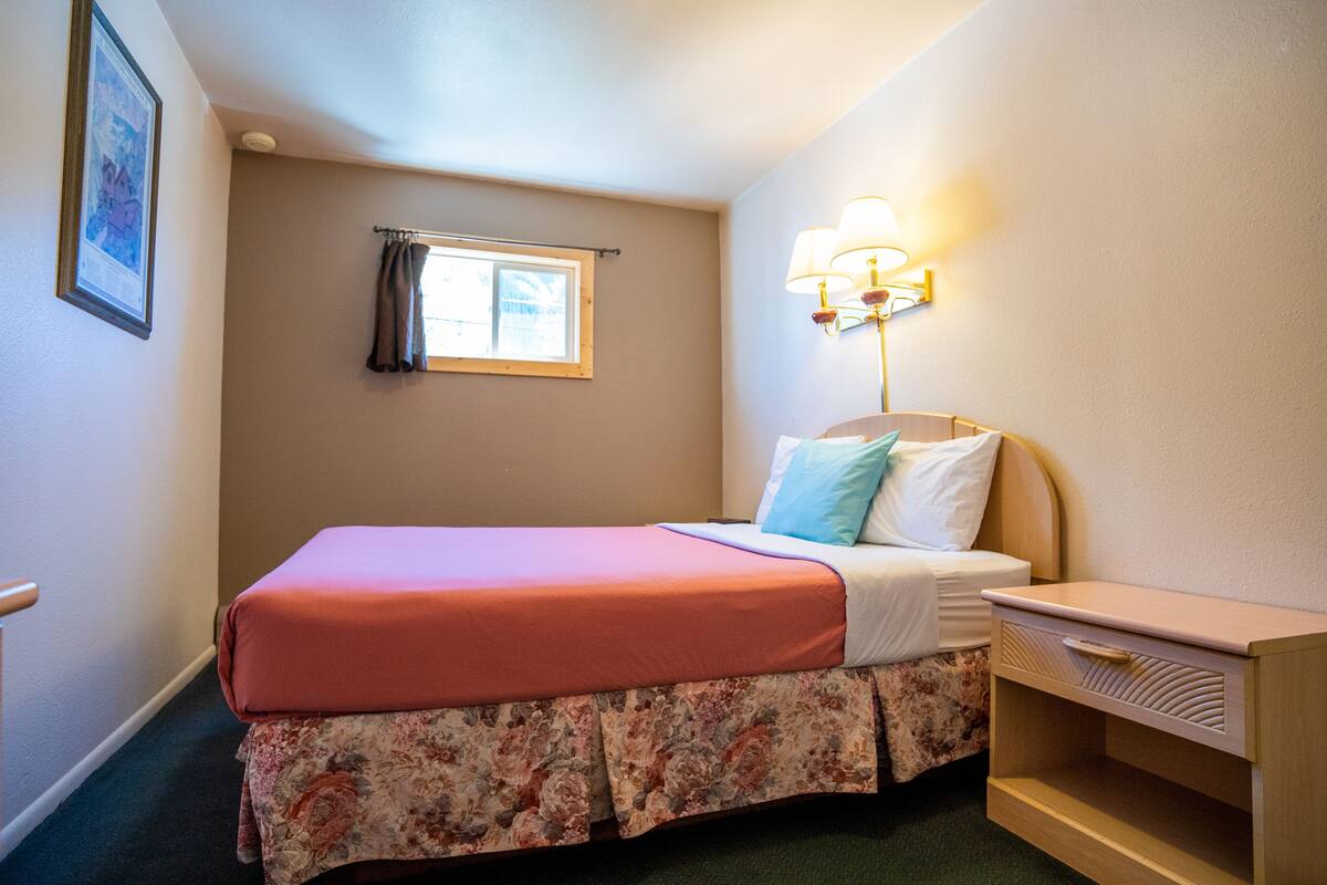 A small, neatly arranged bedroom with a single bed, a nightstand, a lamp, and a window with curtains.